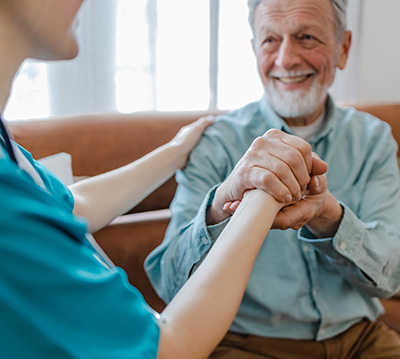 A young female nurse is in the living room with a senior man and holding his hands